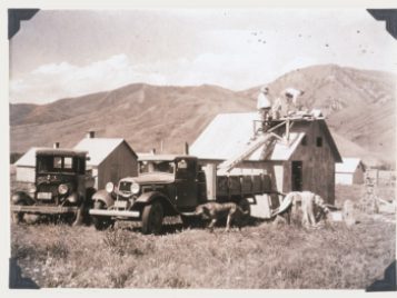 An old, black-and-white photograph of men working on a roof. A 1930s lorry is parked near the building and a dog is running past it.